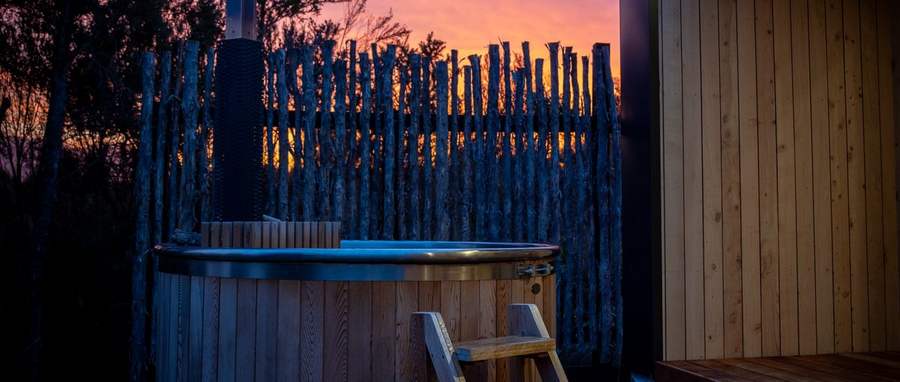 Wood-fired Tubby hot tub at sunset beside a modern cedar home, driftwood fence glowing behind